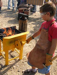 A young boy, dressed in protective leather apron and gloves,is turning the crank on a blower for a small forge.