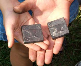 Two hands, one a small child's, the other the dirty hand of an older child.  Each is holding a square "coin" of iron that has been pressed.