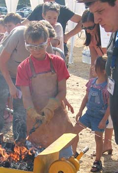 A young boy removing a glowing piece of steel from a coal fire with a pair of tongs.