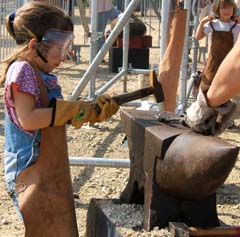 A small girl is using both hands to hammer a hot piece of metal as an adult holds it