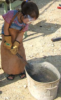 A small girl quenches a hot piece of steel held in a long pair of tongs.  Steam rises from the water.  Her prtective leather apron nearly touches the ground.
