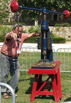 A large man seems small compared to the screw operated mechanical press mounted on a wooden bench.  The press and bench are freshly painted blue and red.