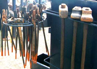 shown is a rack containing perhaps two dozen tongs with various specialized heads, a couple of hammers and three large sledge hammers.