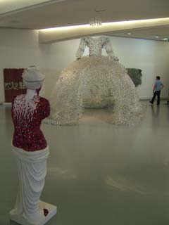 A telve foot tall dress is made of clear plastic bottles.  In the foreground is a female figure covered entirely with what apears to be plastic false-fingernails.