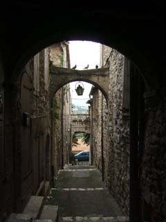 A narrow road heads radially out from the center of town through first a tunnel under the building above, then through a narrow stairway.  A butress between the buildings has pigeons resting on top.  the countryside is visible at the of the long darkened canyon of this walkway.