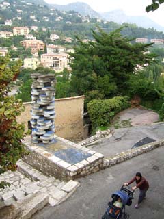 a view from the old western entrance to Vence with a winding road and hills dotted with villas in the distance.  A woman with a head scarf pushes a baby carriage up the road now paved with asphalt.  An abstract sculpture (looks like a stack of concrete puzzle pieces painted blue on the flat side and white on the edges stands ten feet tall on a platform to one side of the road.