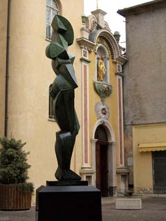 Large bronze sculpture, a cubist abstraction of a woman, stands on a pedestal.  The entracne to the cathedral of Vence is in the background with the Virgin Mary in gold high on a nave above the door to the church.