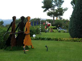 The profile of three women in long dresses are cut out of sheet steel of varying petinas such that the first appears orange while the two in back appear almost like shadows of the first.  The women walk together holding a tube that seems like a rope.  Several other sculptures are visible in the distance.