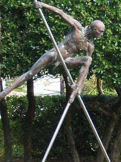 Life-sized bronze man seems to be trying to navigate while suspended several feet above the ground on poles.  He looks foreward with an intense look.