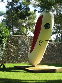 A cylindrical form with rounded ends is standing erect on a low base.  It presents a blue eye and a sad mouth in this view along with a deep gash painted red.