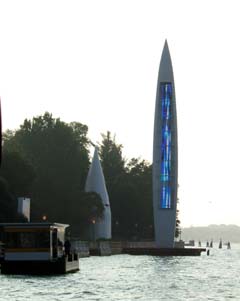 A small yellow water-bus stop sits in front of a large rocket shaped tower.  The tower has a display on one side that has a blue abstracted rendition of water.