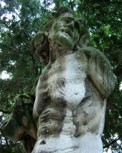 A moss covered statue of an old woman is nearly invisible against the foliage of the trees growing near the Giardini.  She is missing her right forearm and most of her left arm.