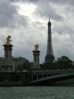 A view of the Seine, a heavily decorated bridge with columns topped with gold colored sculptures and the Eiffel Tower in the background.