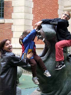 Two children playing on life sized fiberglass sculptures of a Hippopotamus.  A mother is holding the younger child while pretending that there is danger.