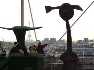 Two abstracted figures face the exhibit with a view of the city of Paris behind.  Other tourists point at sights.