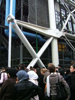 In the foreground is a crowd of people waiting in line.  the background is the huge forgings that make up the external structural elements of the George Pompidous Center.