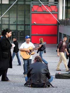 A man stands in a meandering crowd while playing a guitar.  In the background is the distinctive facade of the George Pampidou Center.