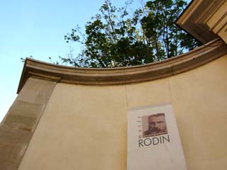 A sign announcing Musee de Rodin hangs on a curved wall.  Glimpses of the sky and folliage contrast with the masonry.