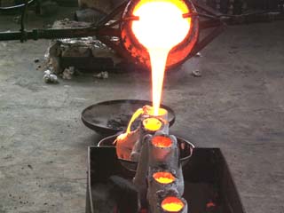 Crusible of bright yellow molten bronze is being poored into molds.  Previously poored molds glow with cooling metal in the foreground.