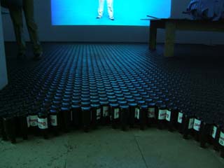 Floor covered in hundreds of inverted "Duvel" beer bottles.  In the foreground the labels can be discerned but beyond the first row, only the bottoms of the bottles can be seen.  In background is a person standing on the bottles and a projected image of a person standing.