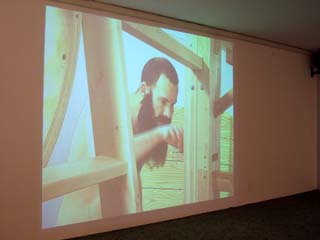 image of the video projection of Guy Ben-Ner interacting with his sculpture.  He has just discovered a small drawer containing tools and hardware for assembling furniture out of the parts of his sculpture.