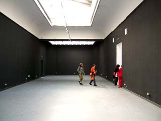 An empty room except for a few people who are leaving.  The walls are black, ceiling white, floor is unfinished concrete.