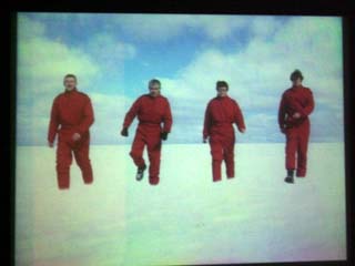 image of a projection screen showing four people dressed in red as they walk in a large field of snow.