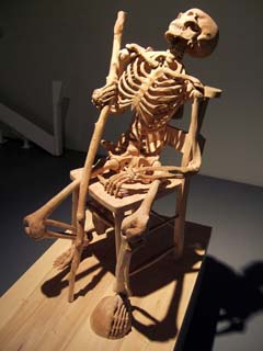 Highly acurate wood carving of a skeleton holding a wlaking stick and sitting in a chair.