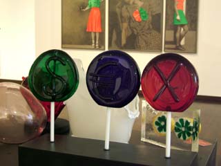 Three glass lollipops are labeled with a dollar sign, a Euro sign, and an X to spell the word SEX.  in the background are other artworks including three images of women.