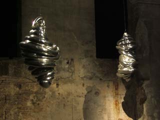 Abstract aluminum objects that look a bit like twisted taffy hang from thin cables in a darkened room.  They shine with reflected light in front of an agind gray masonry wall.