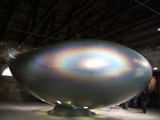 A room sized somewhat teardrop shaped "craft" sits on a stand.  It is painted with a pearlescent grey paint that reflects the light in a series of rainbows.