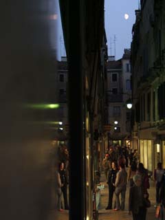 A "wide" street in Venice lined with shops and admiring tourists.  Reflexion in shop window shows same view different angle.  A small patch of sky shows the moon rising.