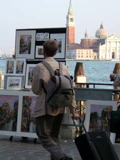 A tourist fresh off the boat walks in front of a street artists display of oil paintings of Venice.  in the background is the Grand Canal and one of many cathedrals.