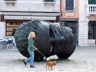 A giant bronze head lies in a stone-paved square in Venice.  Small tufts of grass grow where foot traffic cannot travel.