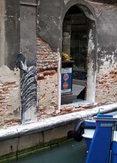 image of a gondolier painted on newsprint and pasted to the crumbling masonry along one of the many canals in Venice.  A boat is visible in the foreground and a Burger King advertisement is visible through an archway in the background.