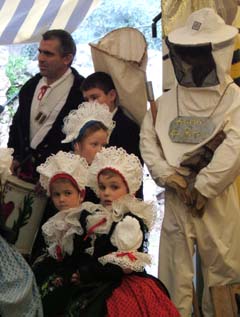 three young girls in full lace stand at the side awaiting their turn to perform.  In the background is a beekeepers geer.