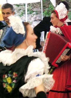 A fuzzy image of a moving dancer in lace and embroidered vest is moving in front of accordian players.