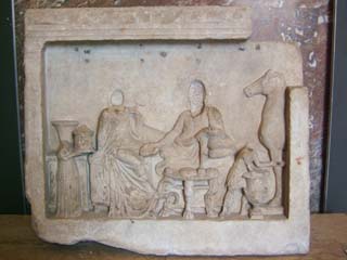 A stone relief depicts two people sitting amongst what must have been a luxurious setting with lots of fabric pieces of furniture and a horse.  The heads are missing and instead are depressions suitable for installing a separate portrait.  Probably a best seller.