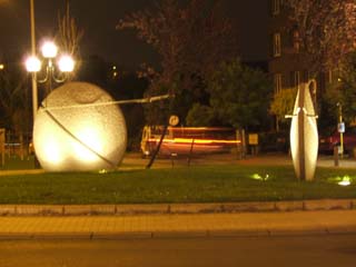 A night photo of two large stone discs each attched to a tree which appears to be pulling the stone.  A streaked image of a car in the background.