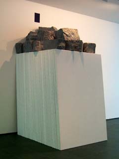 A stack of perhaps fifty presumably blank canvases leans against a wall.  on top of the canvases is a stack of granite blocks.  On the wall above is a short blue stripe.