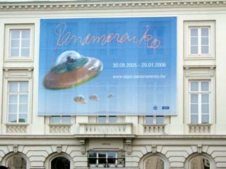 A large blue sign hangs on the side of a building.  Below the name Panamarenko are there flying saucers.