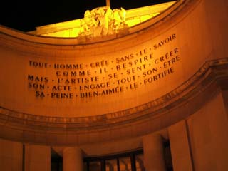 A night photo of a formal facade with an inscription (see text).