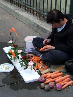 A man carves a carrot in fron of a makeshift display that presents several flowers, fish and birds carved from root vegatables.  A plastic lid contains a few small coins (about two or three euros worth.)