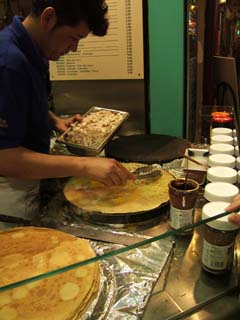 A man is cooking a "crepe complet" on a typical flat fixed creperie griddle.  In the foreground are a stack of crepes and in the background is a menue of offerings and prices.