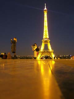 The image shows the Eiffel tower lit up at night.  Their is a reflection of the tower in the smooth stone surface of the terrace.  A woman stands on one side taking a picture and a small family is enjoying the tower.