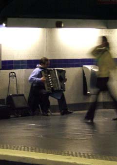 A man plays an accordian in a Metro station with blurred images of passers by.