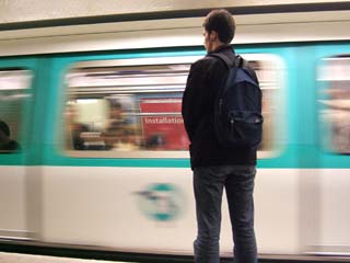 A young man stand as a moving train passes.  A sign visible through the window of the train reads "installation".