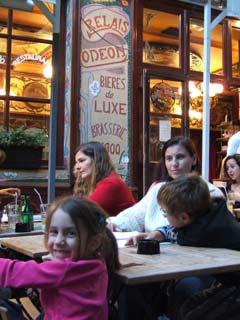 A young girl smiles at the camera while her mother looks inward.  Other people are engaged in conversation iin the background in front of cafe decorated in the Art Nuveau style of the year 1900 or so.