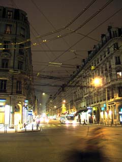 A street is brighltly lit with store fronts and cars but the windows of the buildings above the street level are dark.  A tangle of overhead wires are for the electric buses.