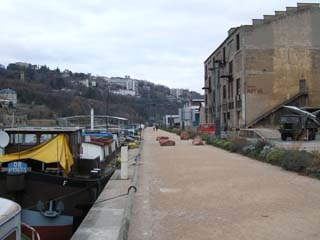 A wide walkway along the river Rhone is lined on one side with travel barges and on the other with wharehouses.  on one side of the image is a missle mounted on a military truck but is only part of the exhibit.